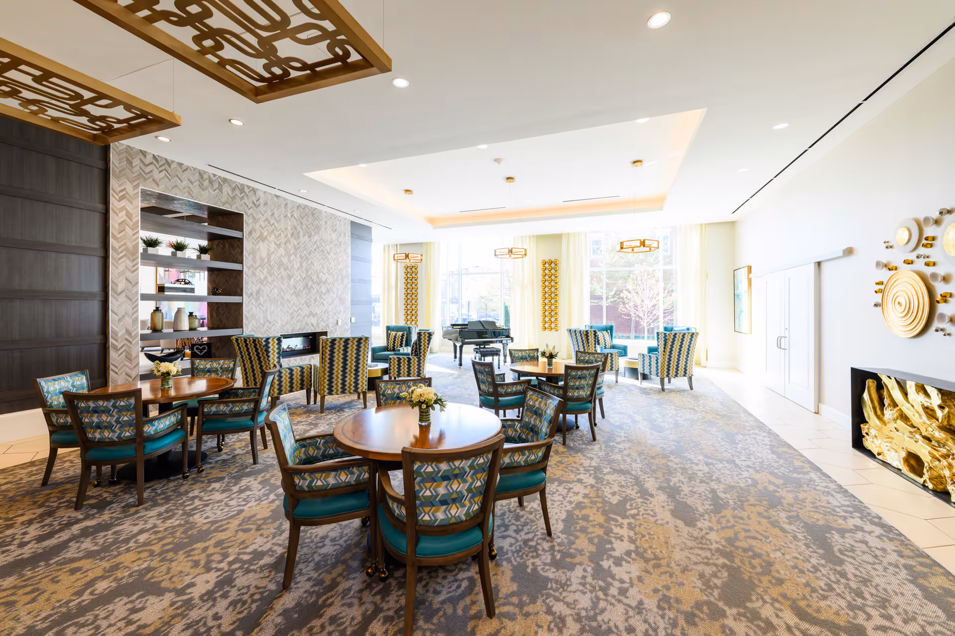 Bright, upscale communal dining and lounge area with round wooden tables, patterned chairs, and a grand piano by large windows.