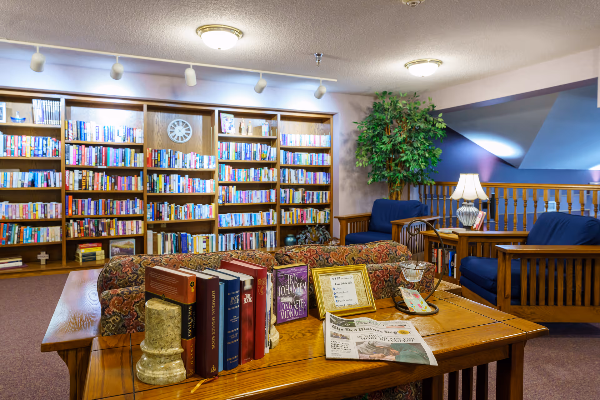 A cozy library or reading room with wooden bookshelves filled with books along the back wall. In the foreground, there is a wooden table with several books, a newspaper, a framed sign, and decorative items. Behind the table are cushioned chairs and a sofa with patterned upholstery. A potted plant and a table lamp are also visible, creating a warm and inviting atmosphere.
