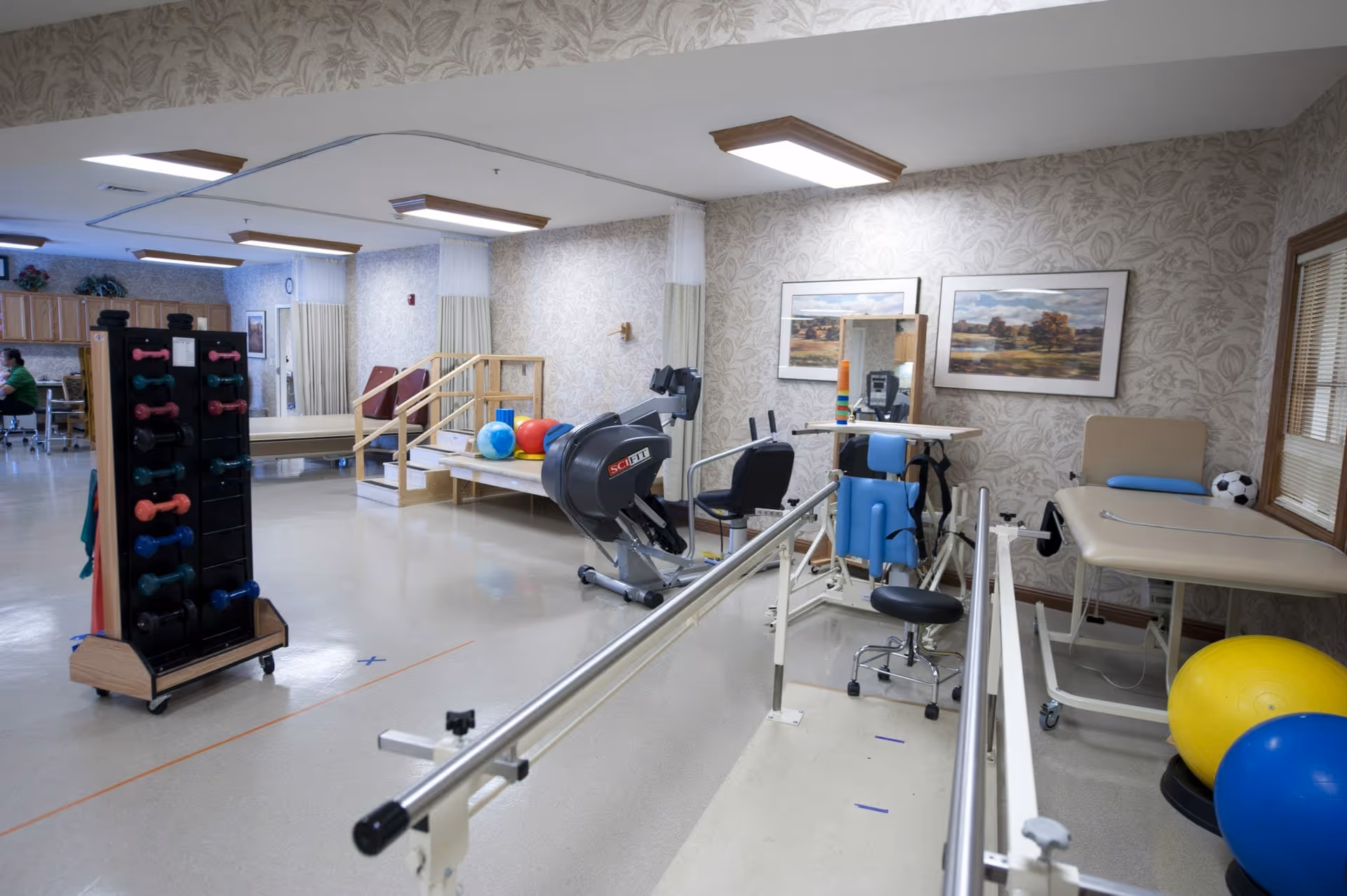 A physical therapy room with parallel bars, exercise equipment, therapy balls, a dumbbell rack, and treatment tables.
