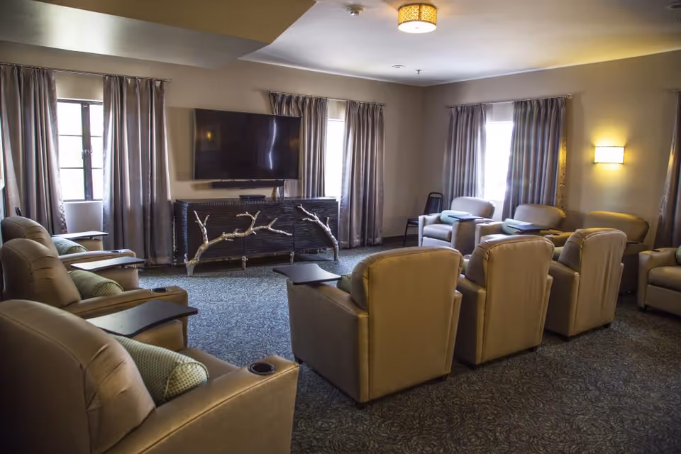 A cozy senior living common area with multiple beige recliner chairs arranged in rows facing a wall-mounted flat screen TV. The room has several windows with gray curtains, a patterned carpet, and soft lighting from wall sconces and a ceiling light fixture.