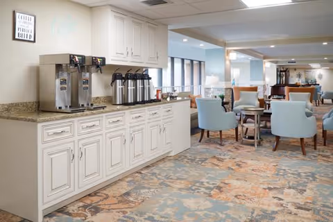 A bright and spacious common area in a retirement community featuring a coffee station with multiple coffee dispensers and creamers on a granite countertop with white cabinetry. The room has light blue upholstered chairs arranged around small wooden tables, large windows letting in natural light, and a patterned carpet with blue, beige, and orange tones.
