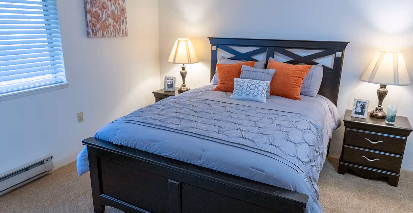 A neatly made bed with a dark wooden frame, adorned with gray and orange pillows and a gray comforter. There are two matching dark wooden nightstands on either side of the bed, each with a lamp and framed photos. A window with blinds and a floral painting are visible on the left wall.