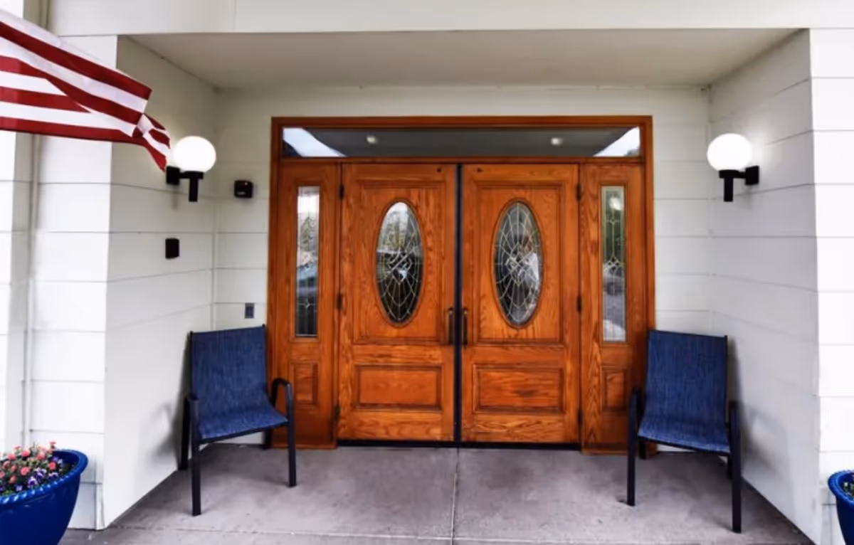 Entrance of a building with double wooden doors featuring oval glass panels. Two blue chairs are placed on either side of the doors, with a blue flower pot containing pink flowers on the left. An American flag is mounted on the left wall, and two round wall lights are positioned on either side of the entrance.
