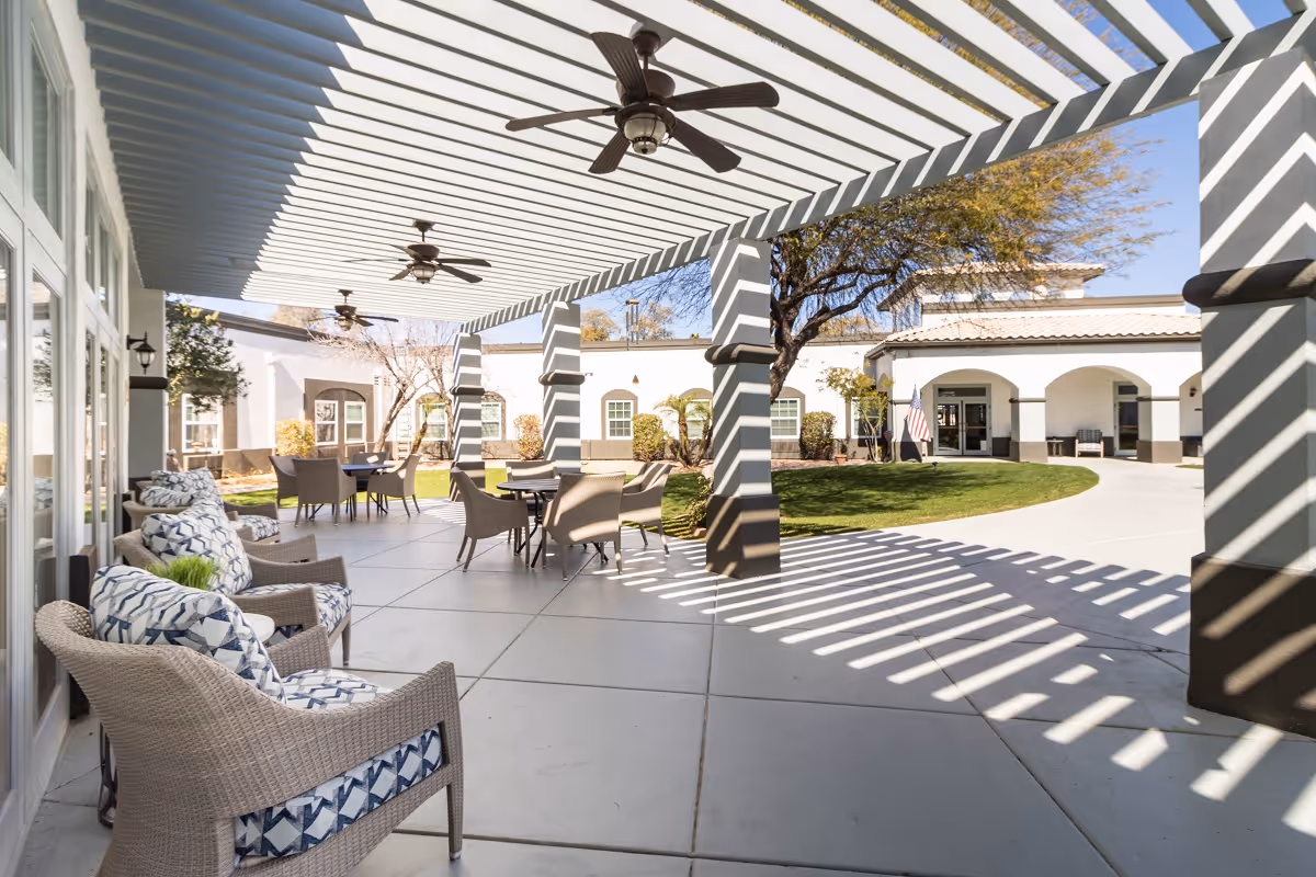 Outdoor covered patio area with wicker chairs and tables under a pergola casting striped shadows. Ceiling fans are mounted on the pergola. The patio overlooks a green lawn and white buildings with arched doorways and windows.