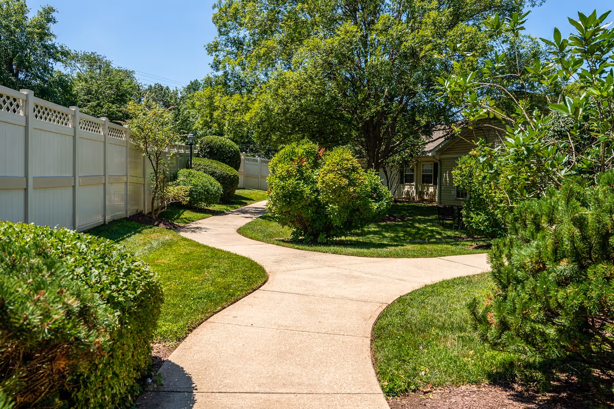A winding concrete pathway through a well-maintained garden area with green grass, bushes, and trees. A white fence runs along the left side, and a building with windows and chairs is partially visible in the background under the shade of a large tree.