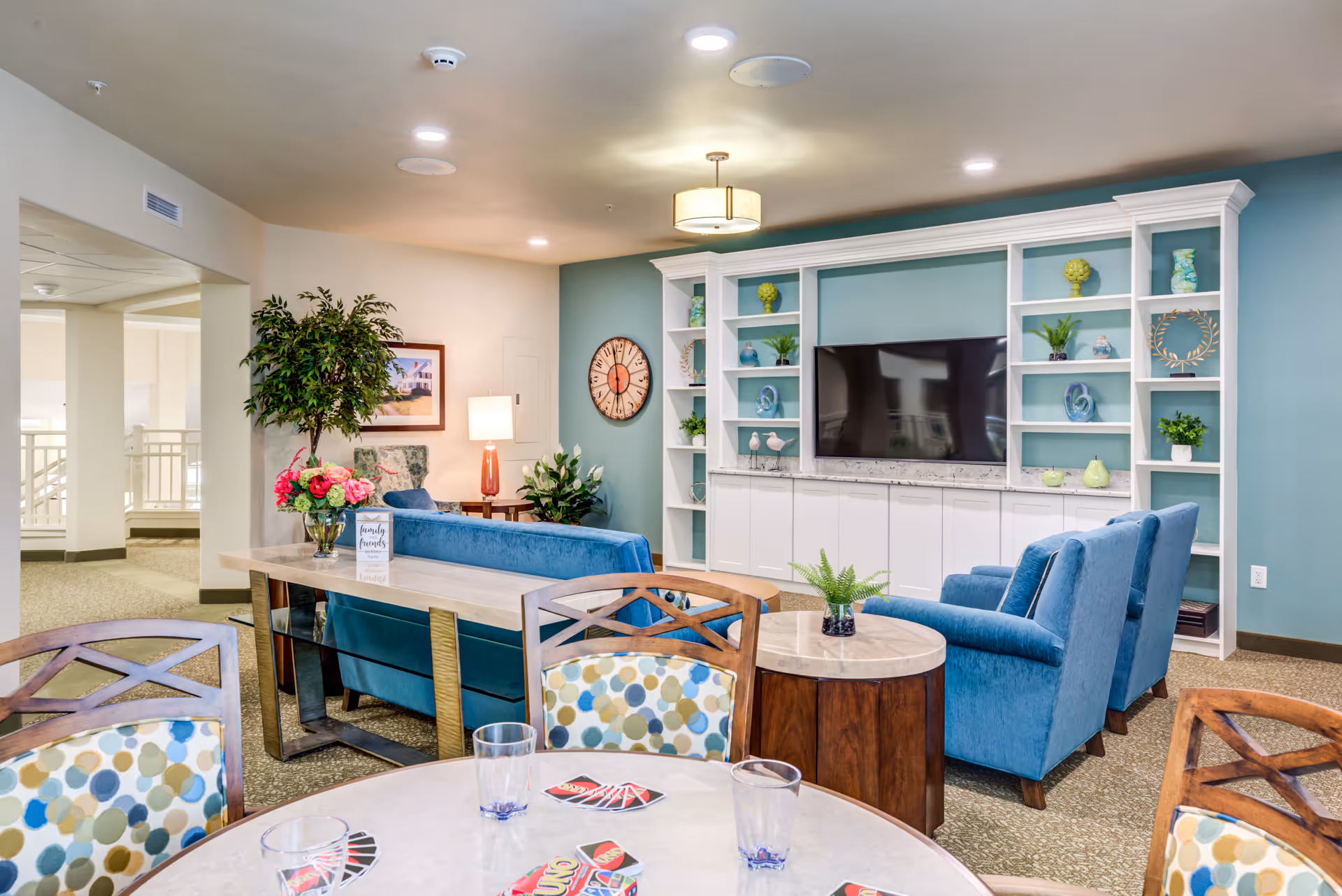 Bright communal living room with blue sofas and armchairs facing a built-in entertainment center and decorative shelving, with a round table in the foreground.