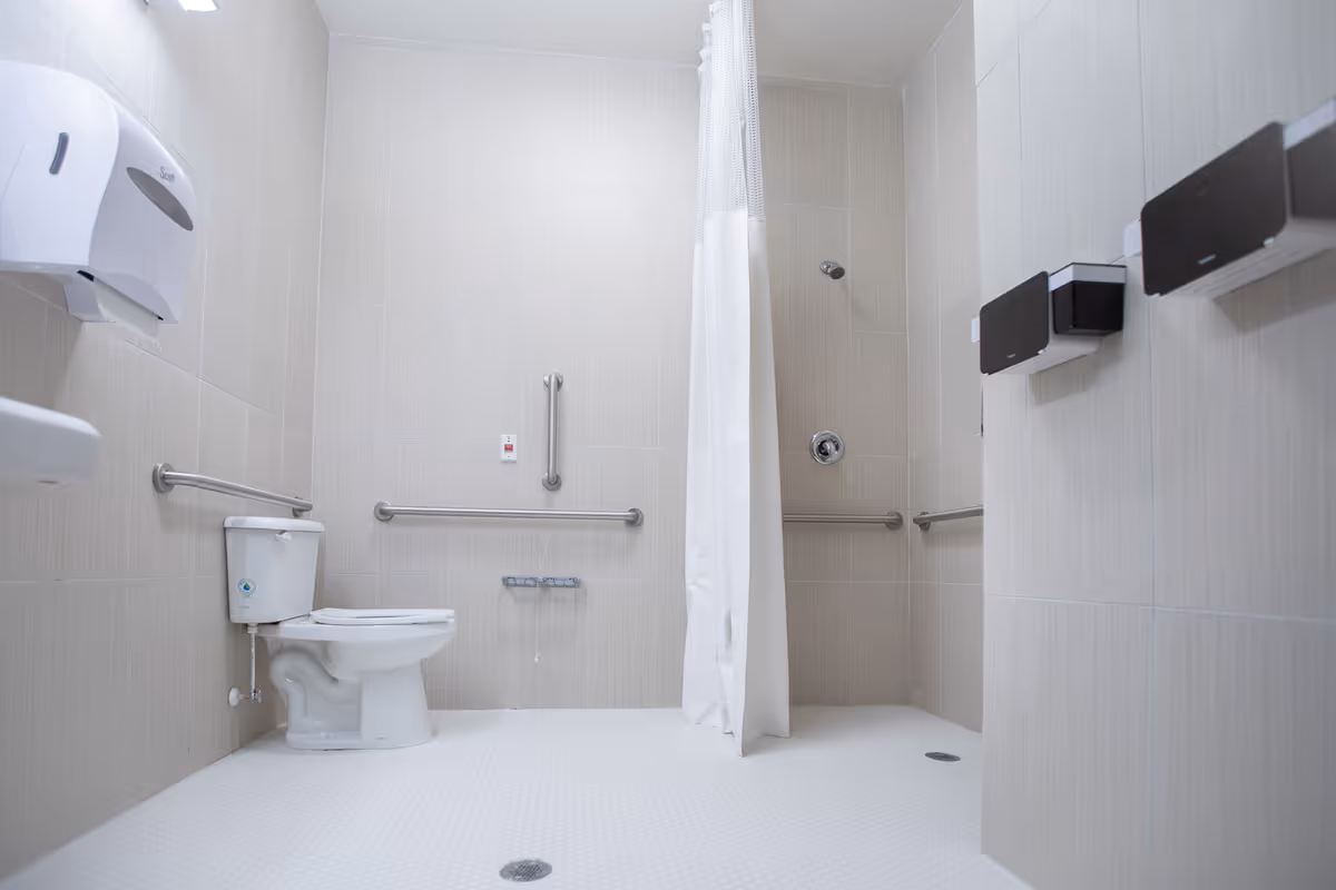 A clean, accessible bathroom with beige tiled walls and white tiled floor. The room features a toilet with grab bars on the walls around it, a shower area with a white curtain, and additional grab bars in the shower. There are also wall-mounted dispensers for soap or paper towels.