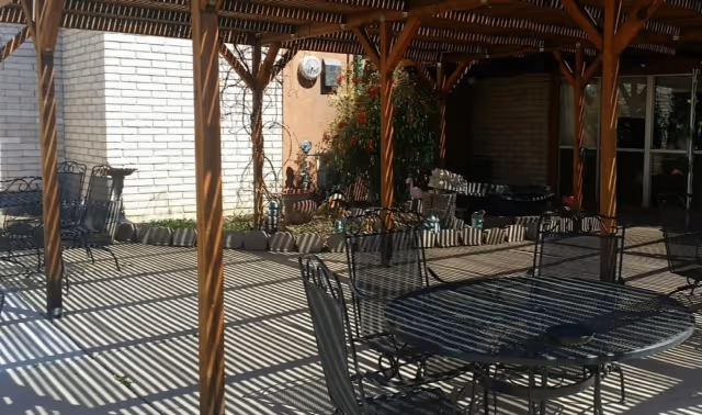 Outdoor patio area with metal tables and chairs under a wooden pergola casting striped shadows on the ground. There are plants and a brick wall in the background.