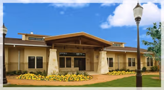 Front entrance of a single-story assisted living building with a covered portico supported by stone pillars, landscaped flower beds and a lamppost under a blue sky.
