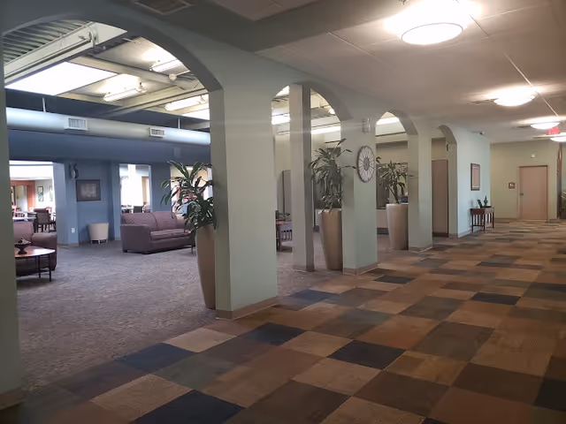 Interior view of a senior living facility hallway with a patterned carpet and several archways leading to a lounge area furnished with sofas and chairs. Large potted plants and a wall clock decorate the space, which is well-lit by ceiling lights.