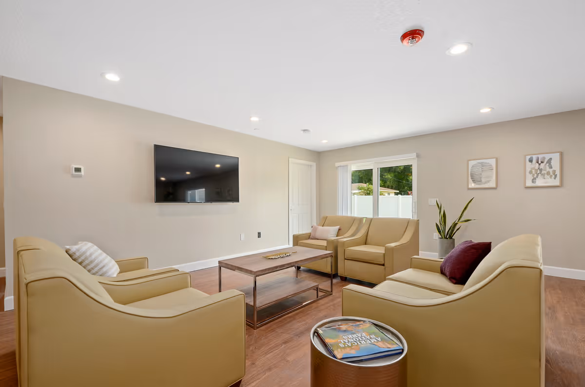 A modern living room with beige walls and wooden flooring featuring four beige armchairs arranged around a rectangular wooden coffee table. A flat-screen TV is mounted on the wall, and there is a sliding glass door with vertical blinds letting in natural light. Two framed abstract artworks hang on the wall, and a potted plant is placed near the door. A small round side table holds a book.
