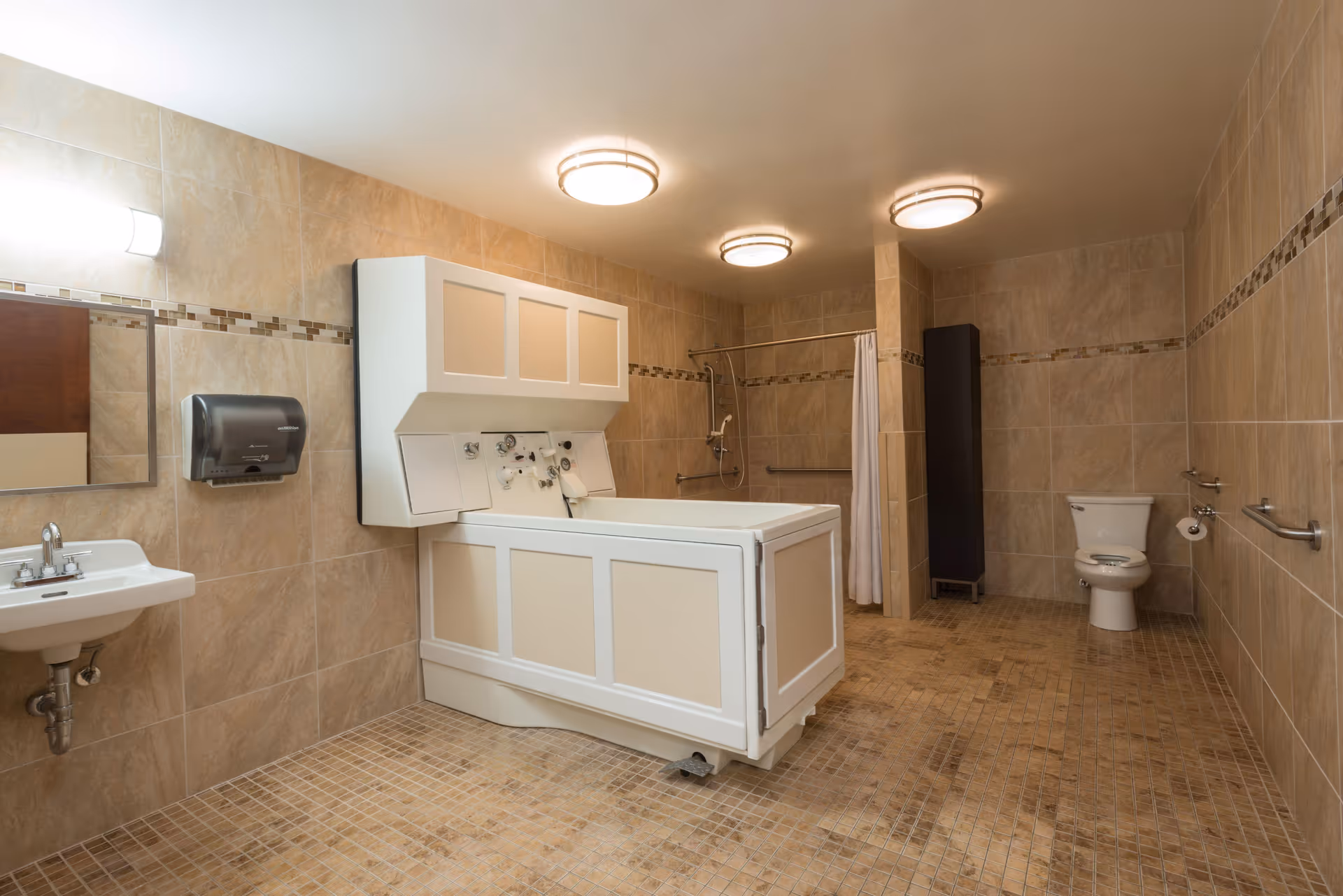 A spacious, well-lit bathroom with beige tiled walls and floor. The room features a white sink with a mirror and paper towel dispenser on the left, a large white accessible bathtub in the center, a shower area with a curtain in the back, and a white toilet on the right. There are grab bars installed near the toilet and shower for safety.