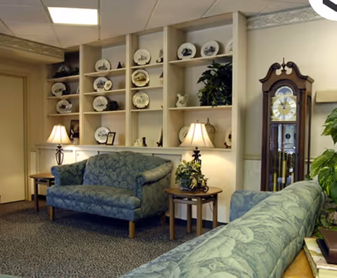 A cozy living room area with two green patterned sofas, two wooden side tables each with a lamp, built-in shelves displaying decorative plates and small plants, and a tall grandfather clock against the wall.