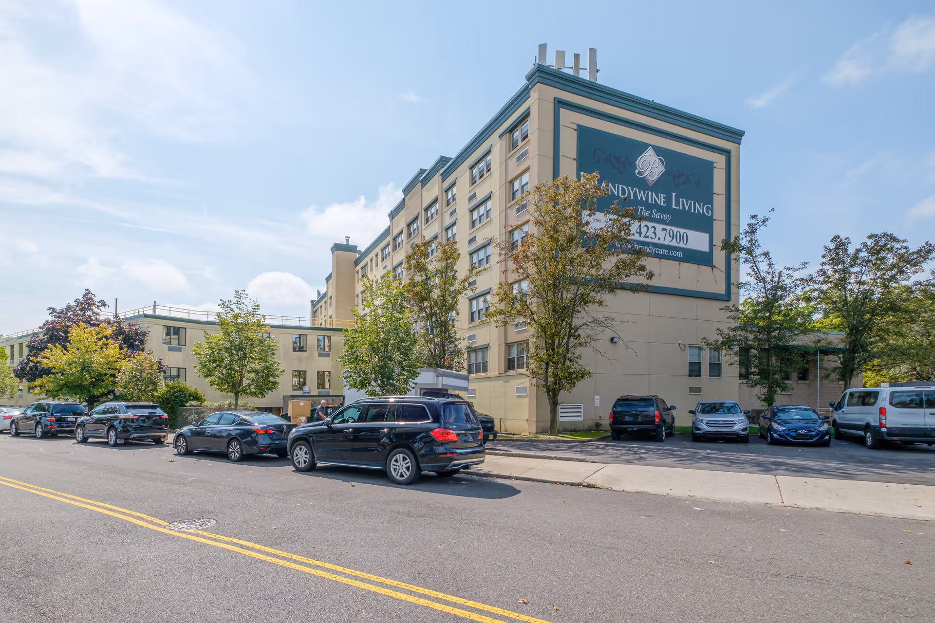 Exterior view of a multi-story senior living facility named Brandywine Living The Savoy, with several parked cars along the street and trees lining the sidewalk under a partly cloudy sky.
