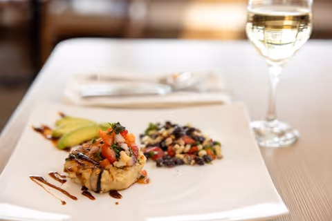A plated gourmet meal featuring a small round serving topped with diced tomatoes and herbs, accompanied by a side of mixed beans and vegetables, garnished with avocado slices and drizzled with a dark sauce, served on a white square plate with a glass of white wine and silverware in the background.