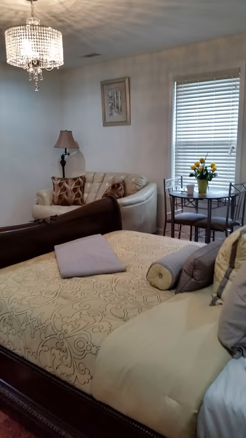 A cozy bedroom featuring a large bed with a beige patterned comforter and multiple pillows. In the background, there is a cream-colored loveseat with decorative pillows, a floor lamp, a framed picture on the wall, and a small round table with two chairs near a window with blinds. A vase with yellow flowers is on the table, and a chandelier hangs from the ceiling.