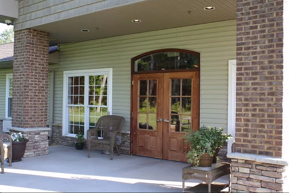 Covered outdoor entrance area of Stonecrest Assisted Living Facility featuring double wooden doors with glass panels, two brick columns, a window with white framing, and wicker chairs with potted plants on a concrete floor.