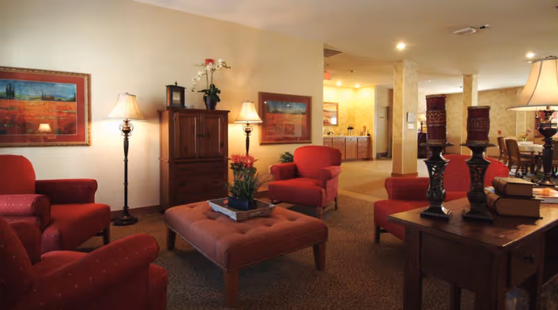 A cozy senior living facility common area with red upholstered armchairs arranged around a large cushioned ottoman. The room features warm lighting from floor and table lamps, framed artwork on the walls, a wooden cabinet with decorative items, and a side table with three large candle holders and stacked books. In the background, there is a dining area with tables and chairs.