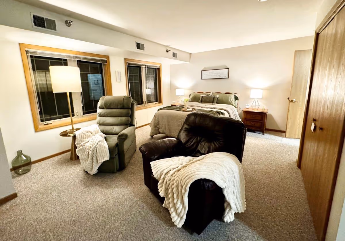 A cozy bedroom with a bed against the far wall, flanked by two nightstands with lamps. In front of the bed are two armchairs, one green and one dark brown, each draped with a white throw blanket. The room has beige carpet, two large windows with wooden frames, and a wooden closet door on the right side.