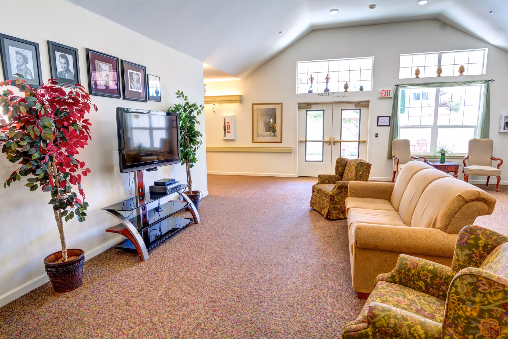 A bright and spacious living room area in a senior living facility with a beige sofa, two patterned armchairs, a flat-screen TV mounted on a stand, framed black and white portraits on the wall, two potted plants, large windows, and double glass doors letting in natural light.