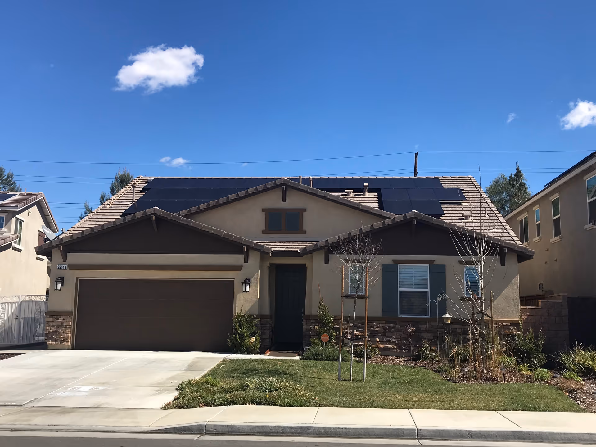 Front exterior view of a single-story house with a two-car garage, solar panels on the roof, a small lawn with young trees, and a clear blue sky with a few clouds.