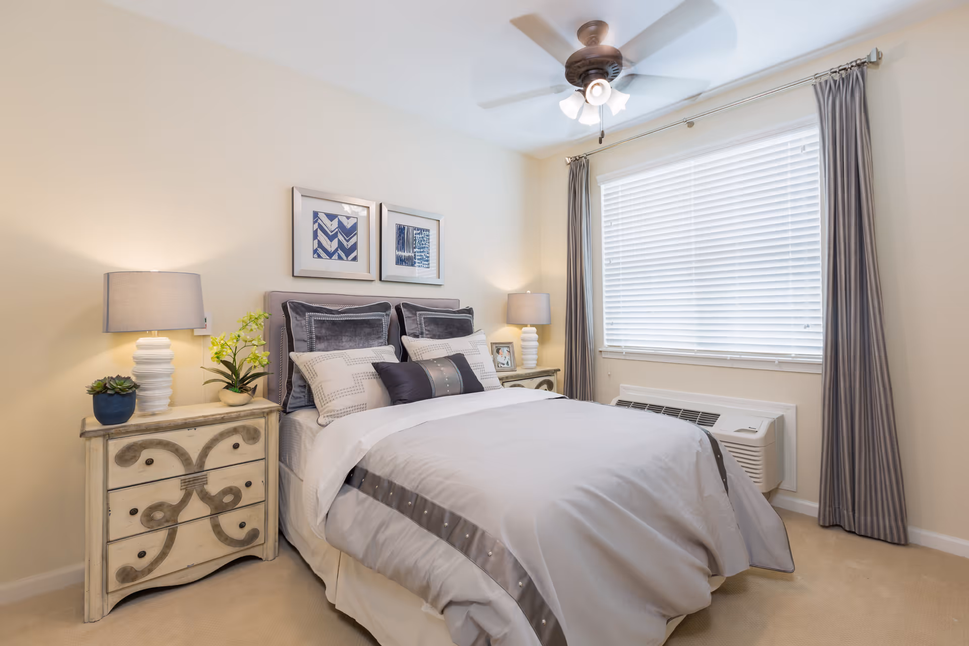 A neatly made bed with gray and white bedding in a bright bedroom. There are two bedside tables with lamps and decorative plants, two framed pictures above the bed, a ceiling fan with lights, and a large window with closed blinds and gray curtains.