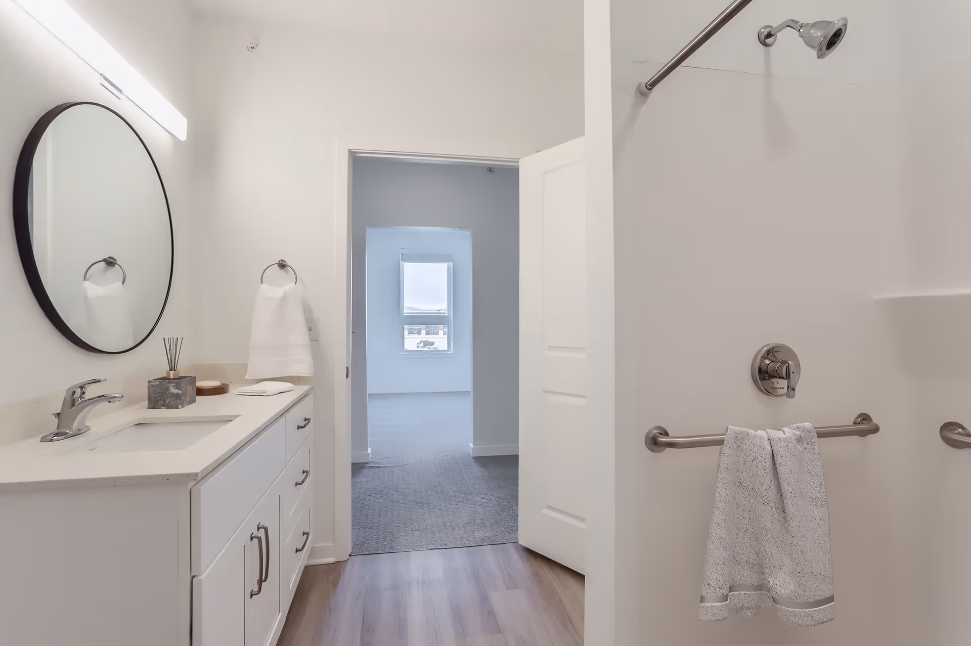 A clean and modern bathroom with a white vanity featuring a sink, faucet, and round mirror above it. A towel ring with a white towel is mounted on the wall next to the vanity. The bathroom has light-colored walls and wood flooring. To the right, there is a shower area with a silver showerhead and a grab bar holding a white towel. An open door leads to a carpeted room with a window in the background.