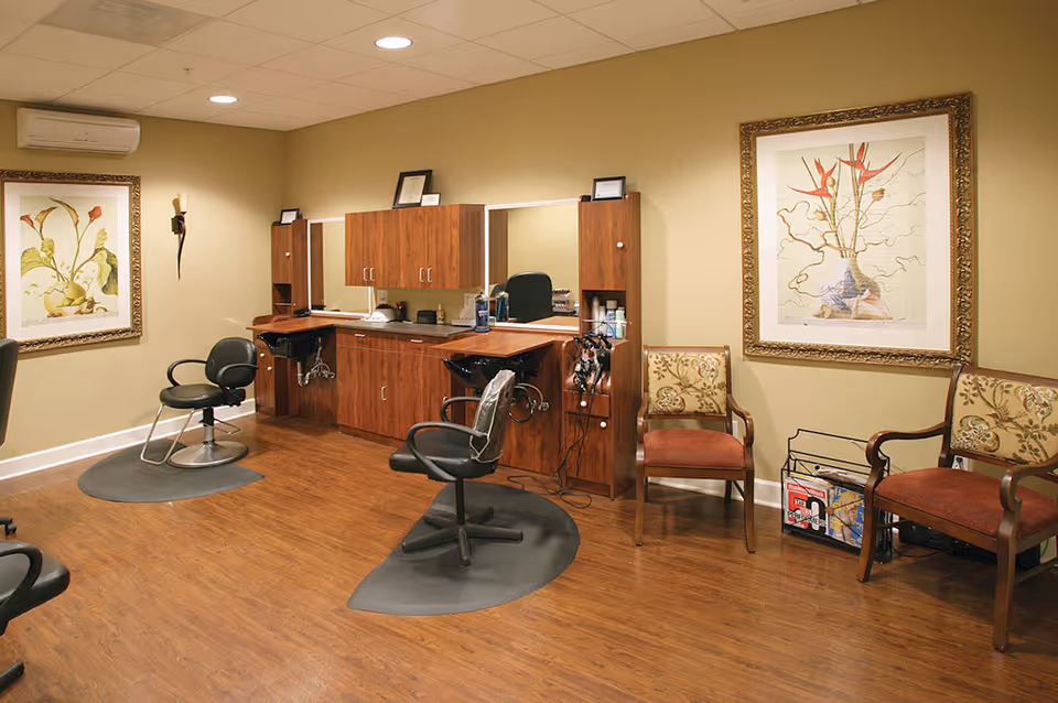 Interior view of a salon area with two black salon chairs on black mats in front of wooden cabinetry with mirrors. The room has wood flooring, beige walls, two framed floral artworks, and three upholstered chairs along the wall with a magazine rack nearby.