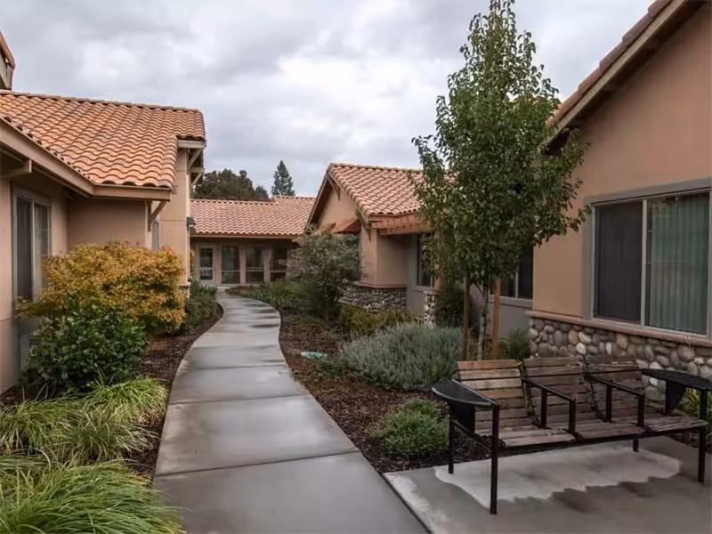 Outdoor walkway between single-story buildings with tan stucco walls and red tile roofs, surrounded by landscaped bushes and trees, with a wooden bench on the right side under a tree.