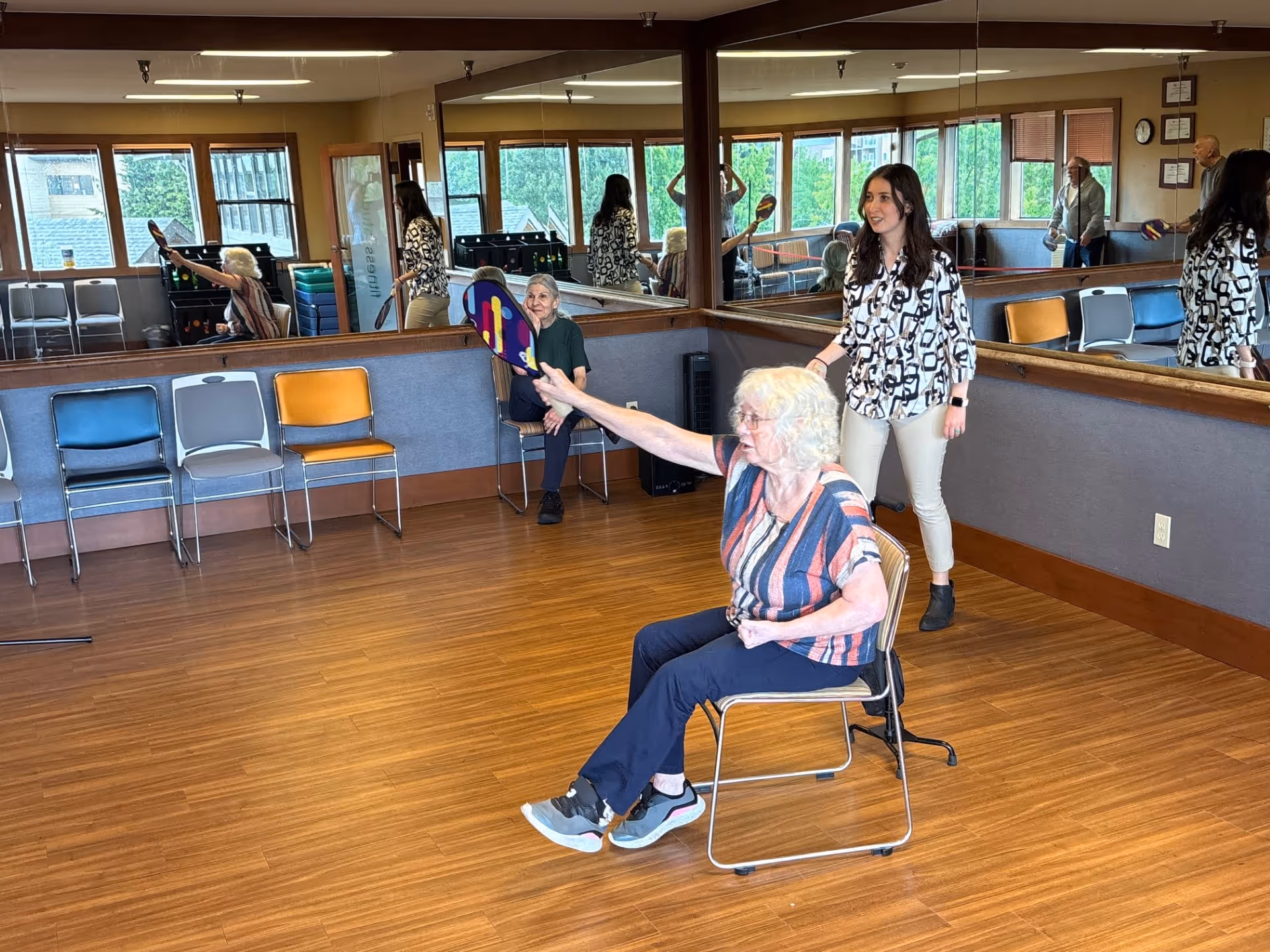 An elderly woman sitting on a chair in a room with wooden floors and large mirrors on the walls, holding a paddle for a game. Another elderly woman is seated in the background, and a younger woman stands nearby, possibly assisting or supervising. Several empty chairs line the wall under windows showing greenery outside.