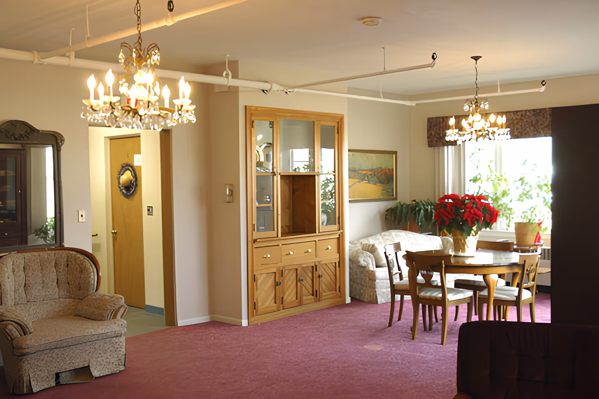 A carpeted communal living and dining area with chandeliers, a wooden dining table and chairs, sofas, a display cabinet and potted plants by a window.