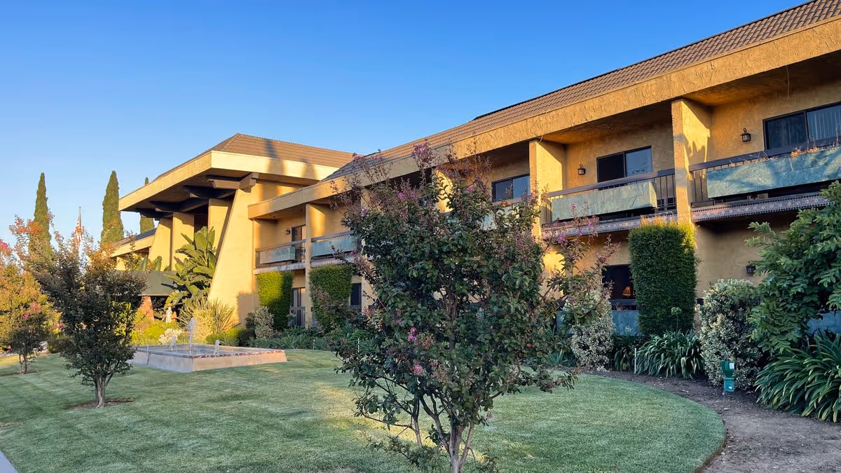 Exterior courtyard and front of a two-story retirement hotel with balconies, lawn, trees, and a small fountain.