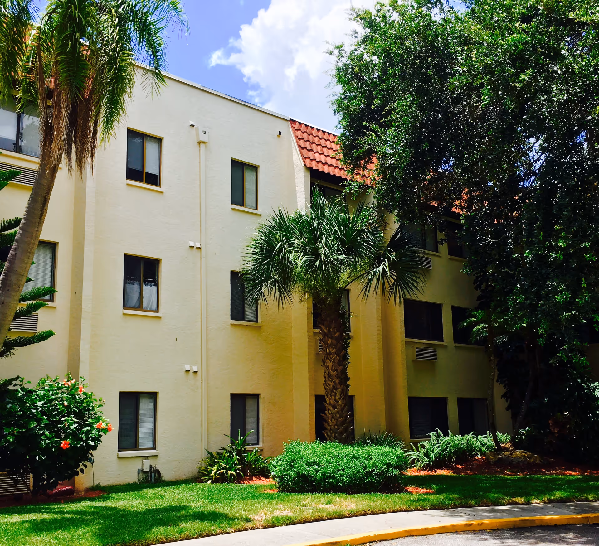 Exterior view of a pale three-story residential building with a red tile roof, palm trees, and landscaped grounds.