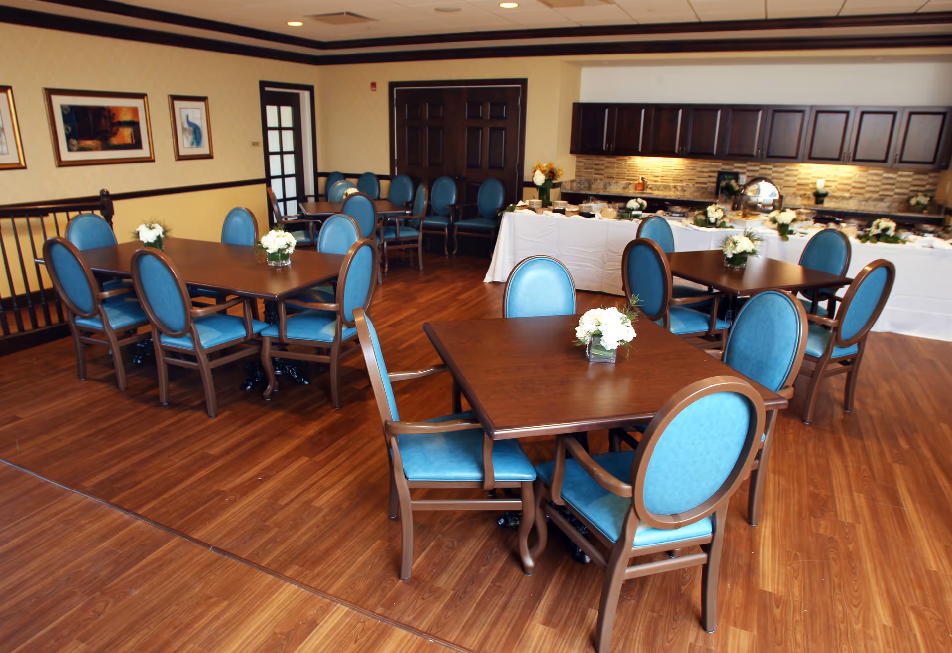 A dining room with several wooden tables and blue upholstered chairs. Each table has a small floral centerpiece. In the background, there is a buffet table covered with a white tablecloth and various dishes, set against a wall with dark wooden cabinets and a tiled backsplash. The room has wooden flooring and framed artwork on the walls.