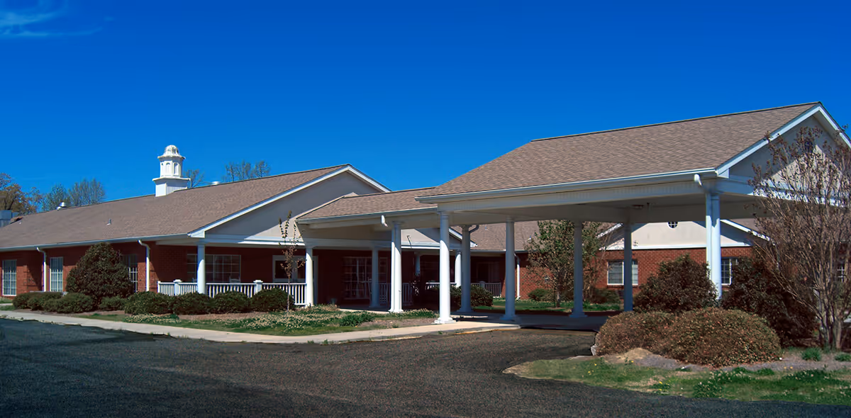 Exterior view of a single-story brick building with a covered entrance supported by white columns, surrounded by shrubs and trees under a clear blue sky.
