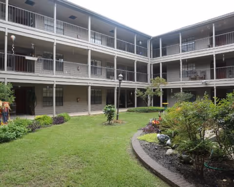 A three-story building surrounding a green courtyard with grass, shrubs, and small trees. The building has balconies with railings on each floor overlooking the courtyard.