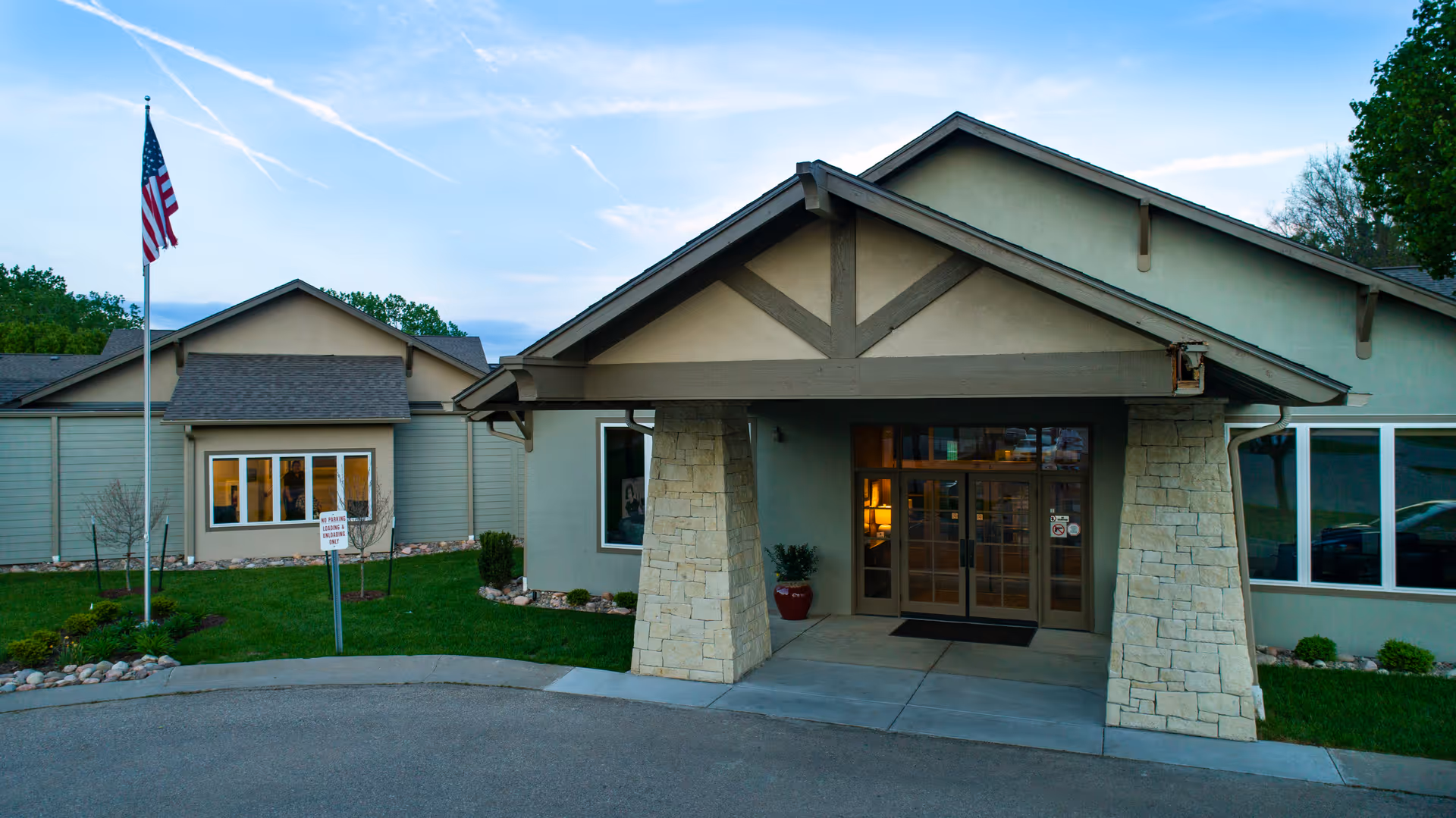 Exterior front entrance of a single-story assisted living facility with a covered porch supported by stone pillars, an American flag on a flagpole, and well-maintained landscaping including grass and small bushes.