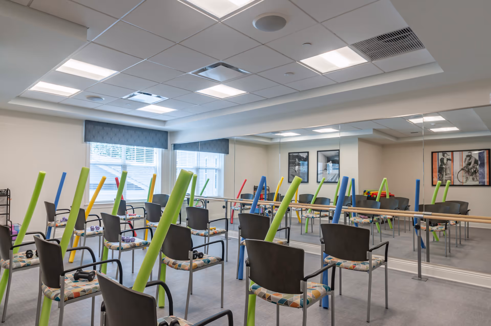 A bright exercise room with chairs arranged in rows, each chair holding colorful foam noodles. The room has large windows with blue valances, a mirrored wall with a ballet barre, and framed black and white photos on the walls.