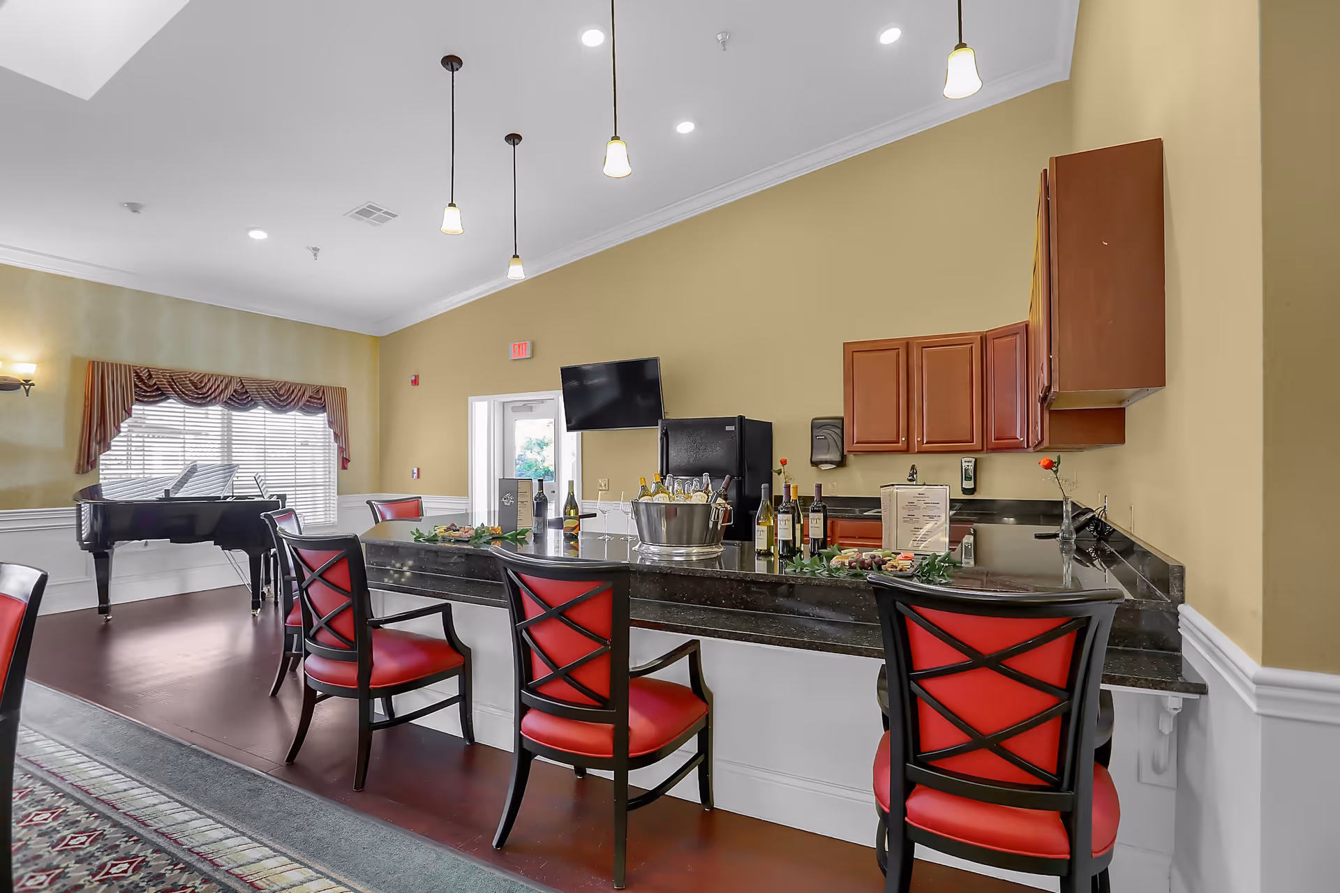 Interior view of a senior living facility lounge area with a black granite countertop bar, red cushioned chairs, wooden cabinets, a black refrigerator, and a black grand piano near a window with blinds and valance curtains. The room has beige walls, pendant lights hanging from the ceiling, and a wall-mounted TV.