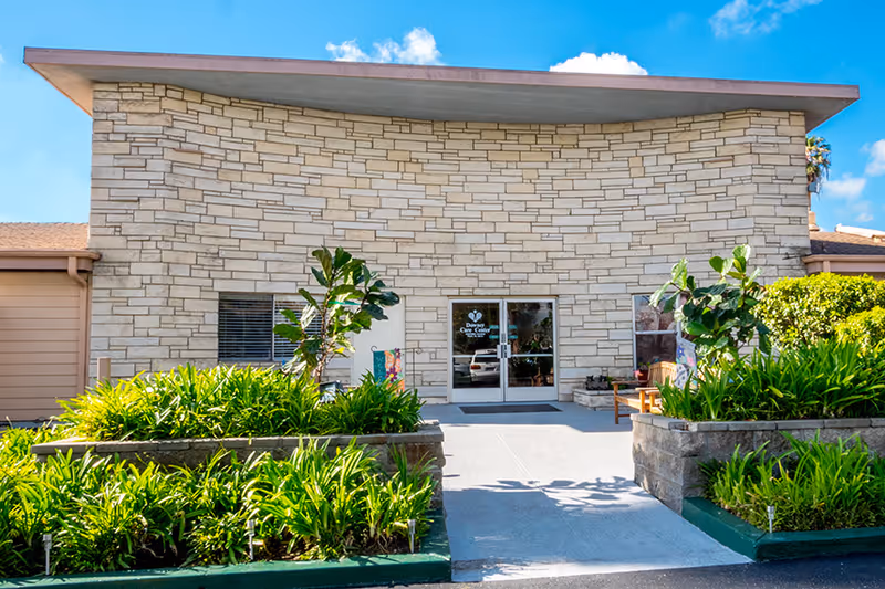 Front exterior view of a building with a stone facade and a flat roof under a blue sky with some clouds. There are green plants and shrubs in raised garden beds on either side of a concrete walkway leading to glass double doors entrance. A bench and some decorations are visible near the entrance.