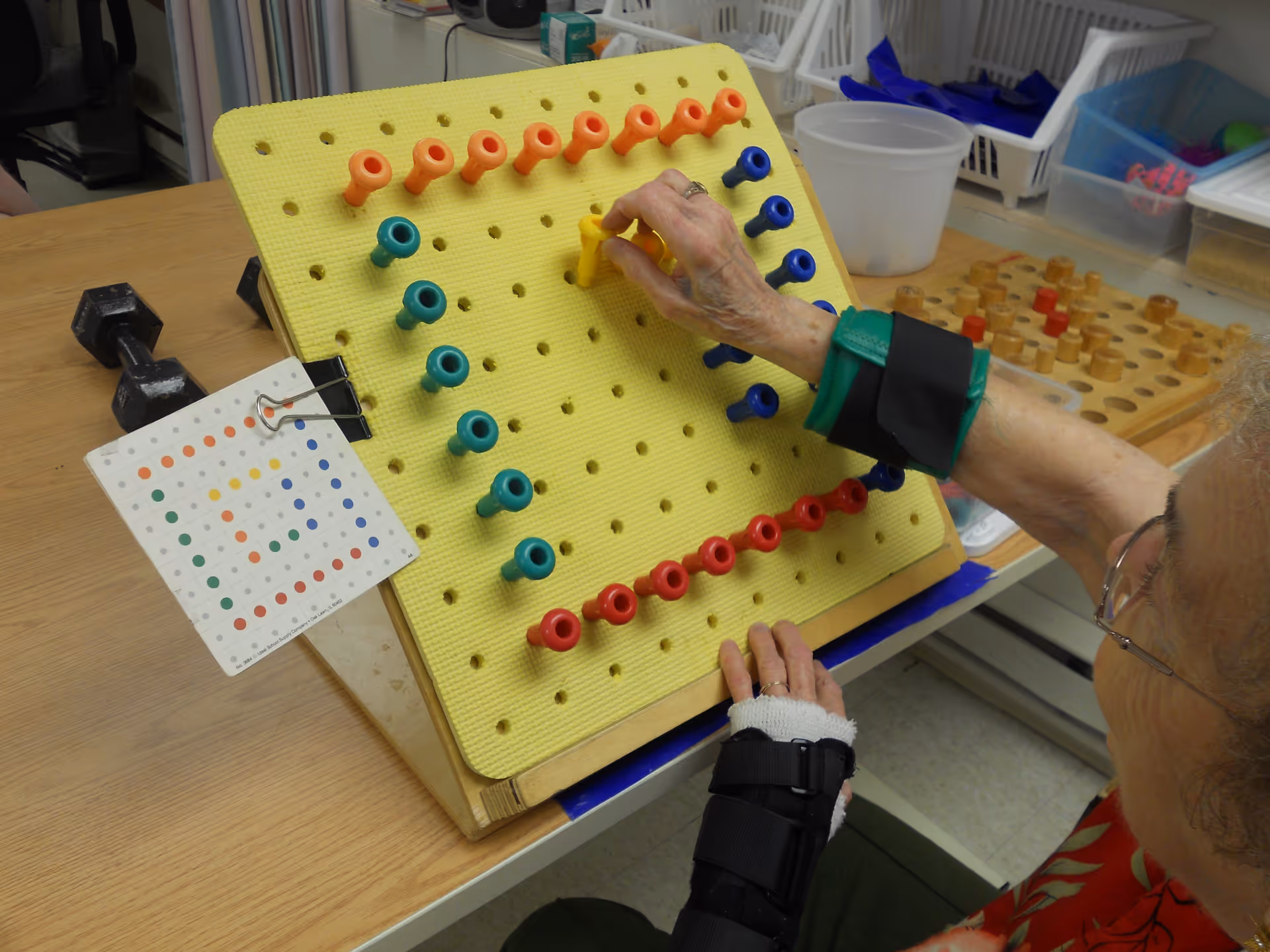 An elderly person wearing wrist and hand braces is engaging in a pegboard activity with colored pegs on a yellow board, likely for hand therapy or cognitive exercise, at a table with other therapy tools nearby.