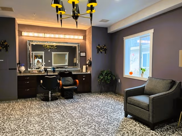 A well-lit room with a large mirror above a counter with two salon chairs in front. The walls are painted purple, and there is a patterned carpet on the floor. A gray armchair is positioned near a window with a small plant and a pumpkin on the windowsill. A chandelier with yellow lampshades hangs from the ceiling.