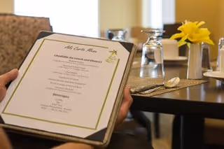 A person holding a menu titled 'À la Carte Menu' in a dining area with a table set with glasses, silverware, and a small vase with a yellow flower.