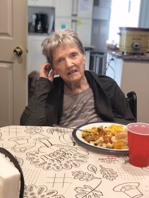 An elderly woman sitting at a dining table with a plate of food and a red cup in front of her. The background shows a kitchen area with appliances and cabinets.