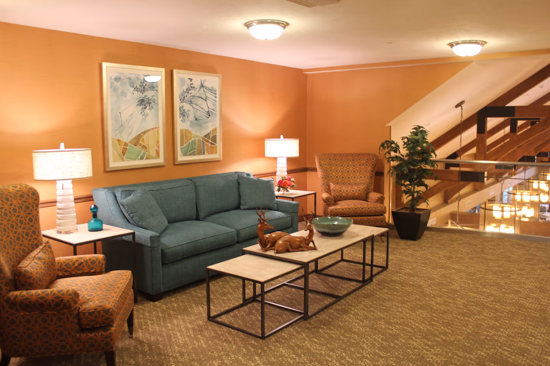 A cozy living room area with a teal sofa, two patterned armchairs, two side tables with lamps, a coffee table with decorative items, framed artwork on the wall, a potted plant, and a staircase with wooden railings in the background.