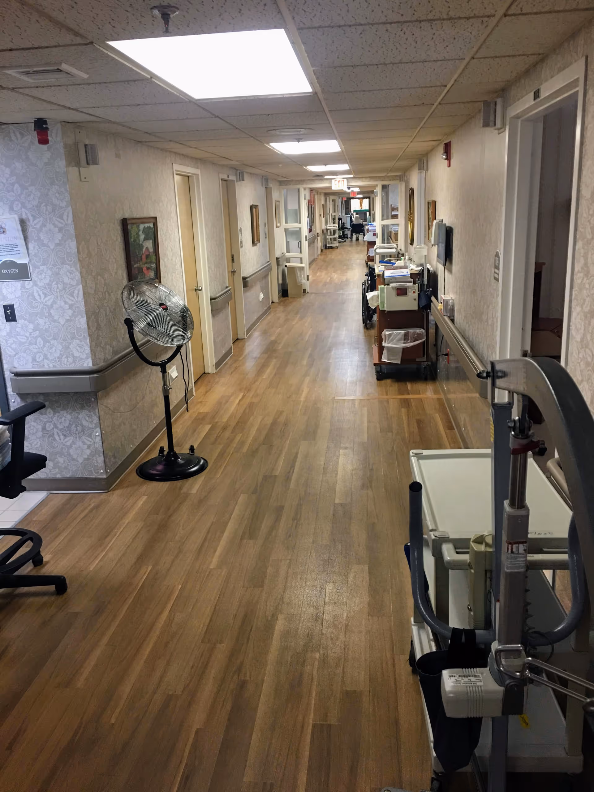Long indoor hallway in a senior living facility with wood-look flooring, handrails, doors and medical carts lining the corridor.