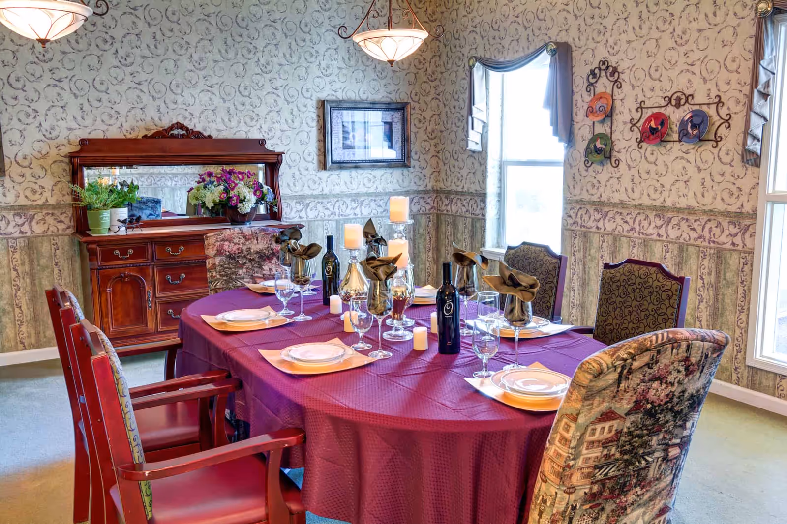 A dining room with a round table covered in a purple tablecloth, set with plates, glasses, napkins, and wine bottles. The room features patterned wallpaper, a wooden sideboard with a mirror and flower arrangements, and several upholstered chairs around the table. Two windows with draped curtains allow natural light into the room.