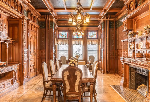 An ornate dining room with a long wooden dining table surrounded by eight upholstered chairs. The room features intricate wood paneling on the walls, a decorative wooden ceiling with beams, and a chandelier with multiple lights hanging above the table. There are three large windows with stained glass at the top and lace curtains, and a fireplace with detailed wood carvings and decorative items on the mantel.
