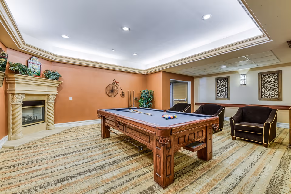 A well-lit recreational room with a pool table in the center, a fireplace with decorative plants on the left, three dark upholstered armchairs on the right, and wall decorations including a vintage bicycle sculpture and framed art pieces.