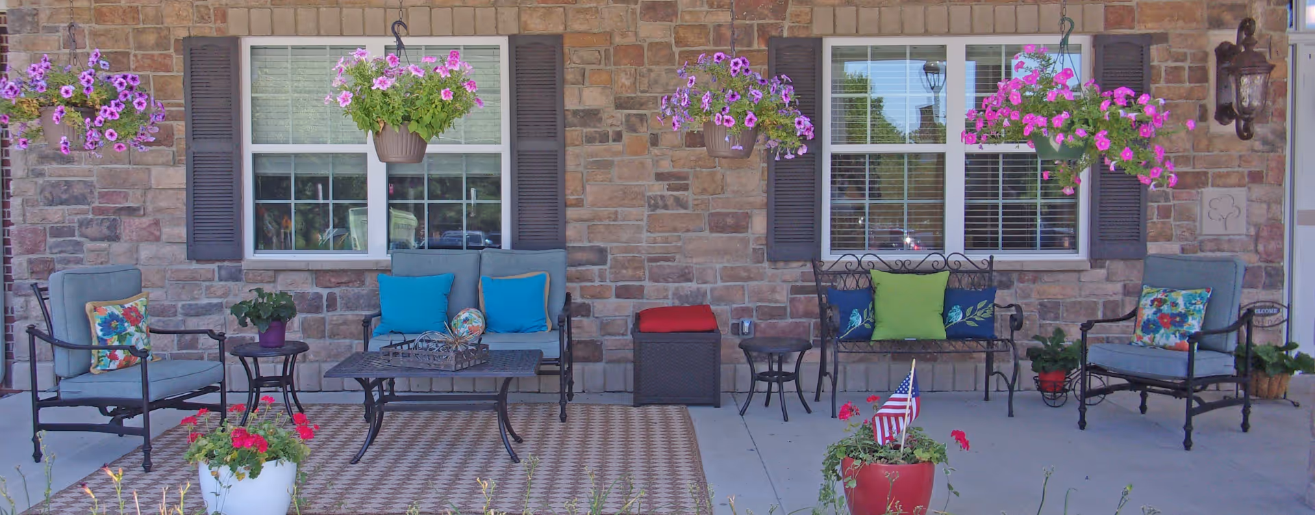 Outdoor patio area with cushioned chairs, a loveseat, a small table, and decorative pillows. Hanging flower pots with purple and pink flowers are suspended above. The background features a brick wall with two windows and a wall-mounted lantern light fixture. Potted plants and an American flag in a flower pot are placed on the concrete floor.